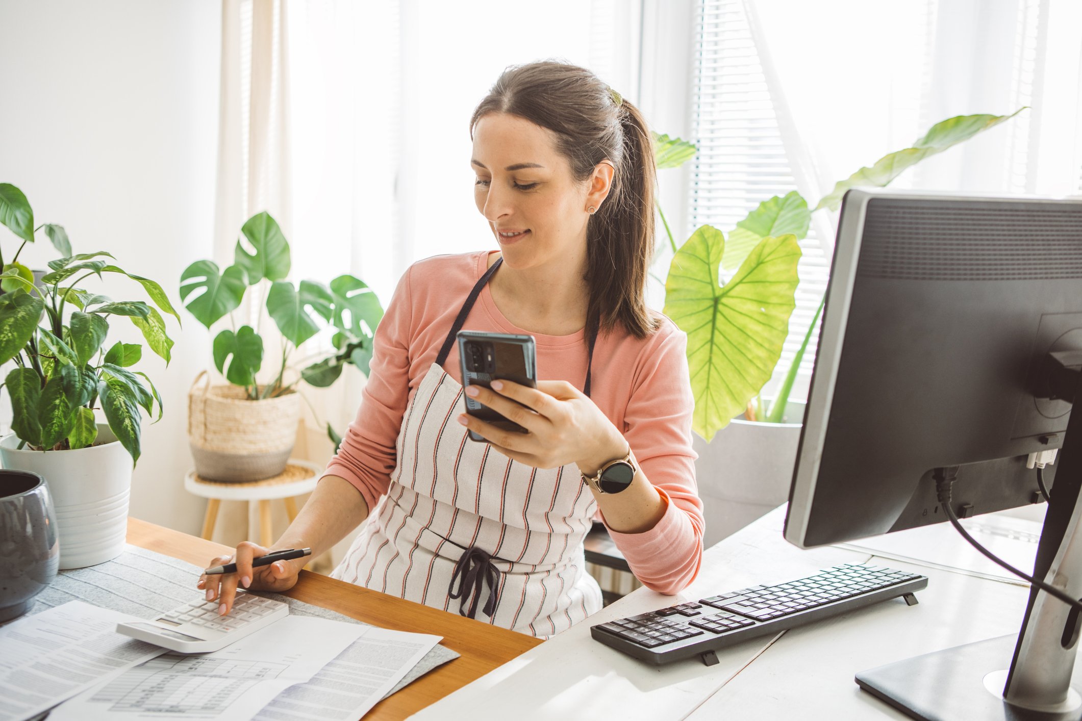 Woman wearing a striped apron punching in numbers into a calculator while holding her smartphone and sitting at her desk with papers and a computer