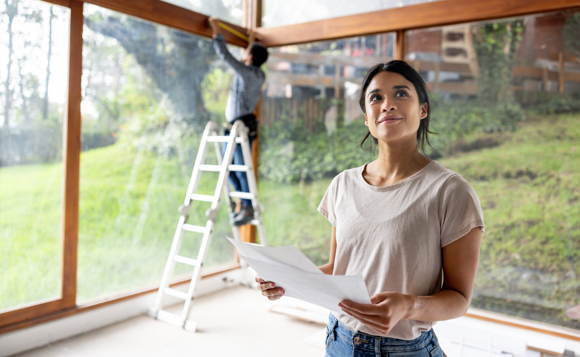 Woman looking up from reading building plans with a person in the background on a ladder measuring the windows inside a house
